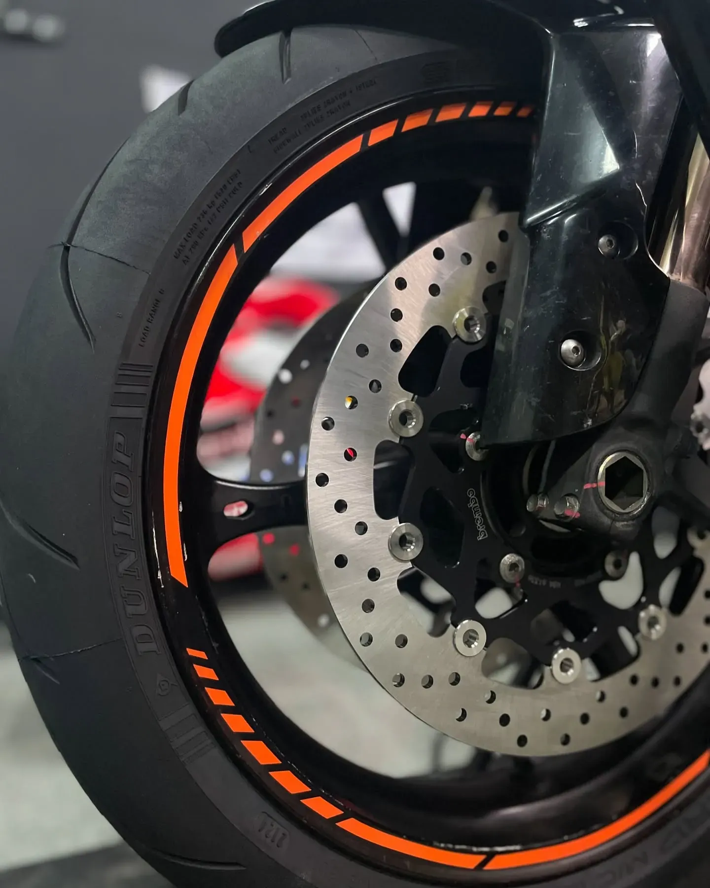 Detalle del freno delantero: disco perforado, pinza y neumático Dunlop con vinilo naranja en la llanta
