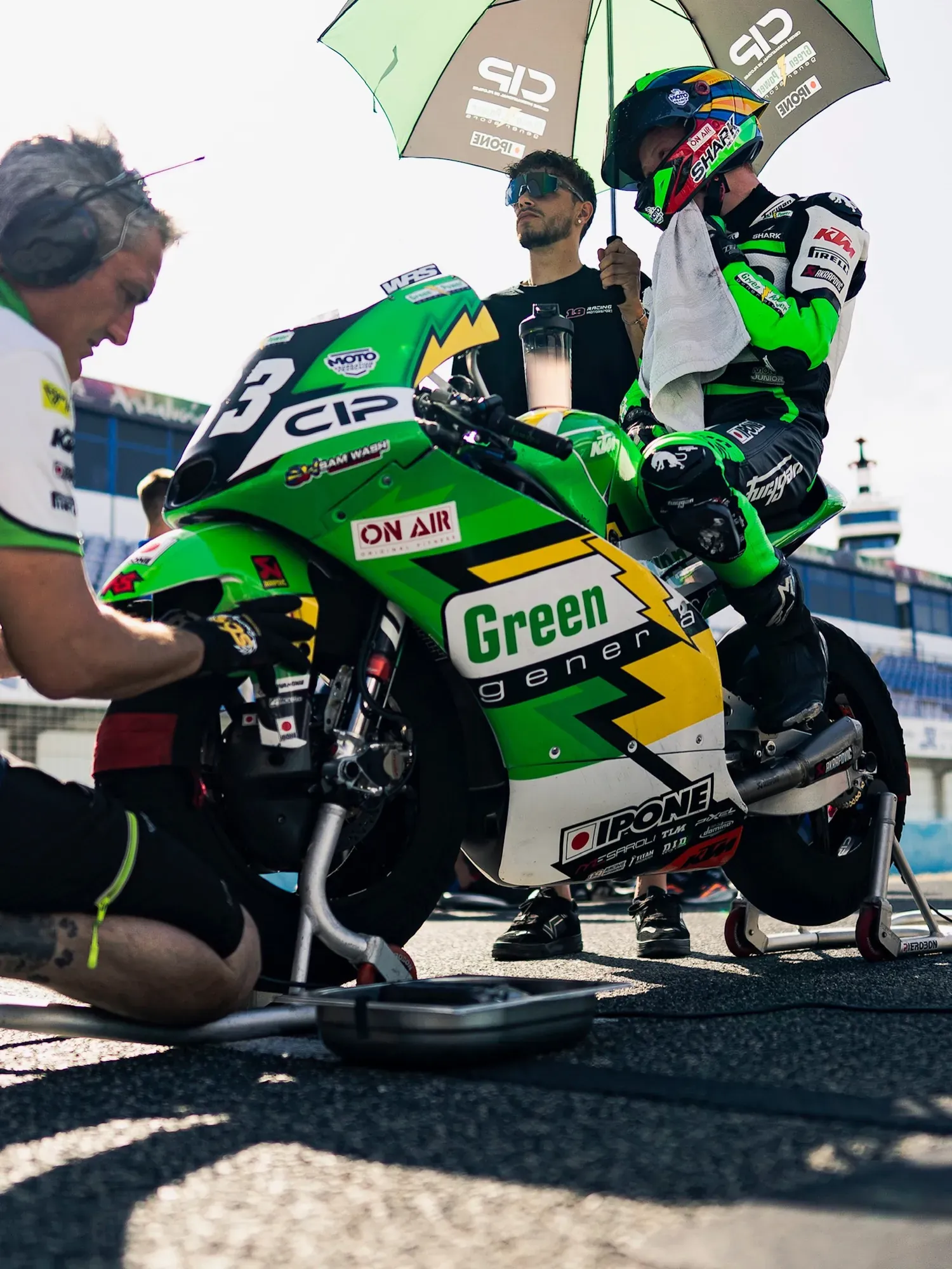 Mecánico ajustando una moto de competición verde en la parrilla de salida, con el piloto subido bajo el paraguas del equipo