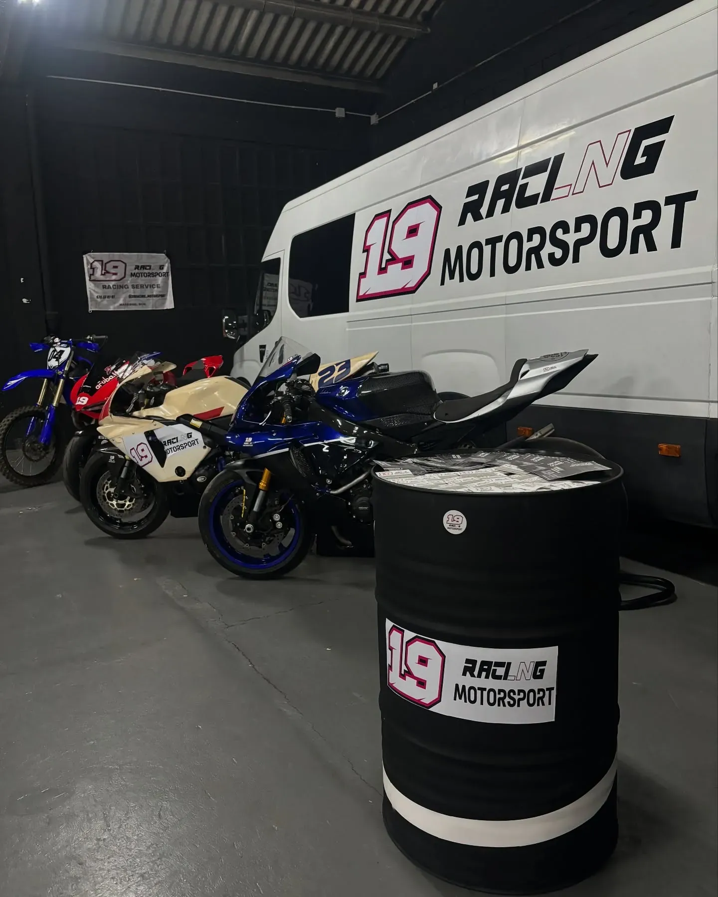 Interior del taller con motos de competición alineadas junto a la furgoneta y un bidón con el logo de 19 Racing Motorsport